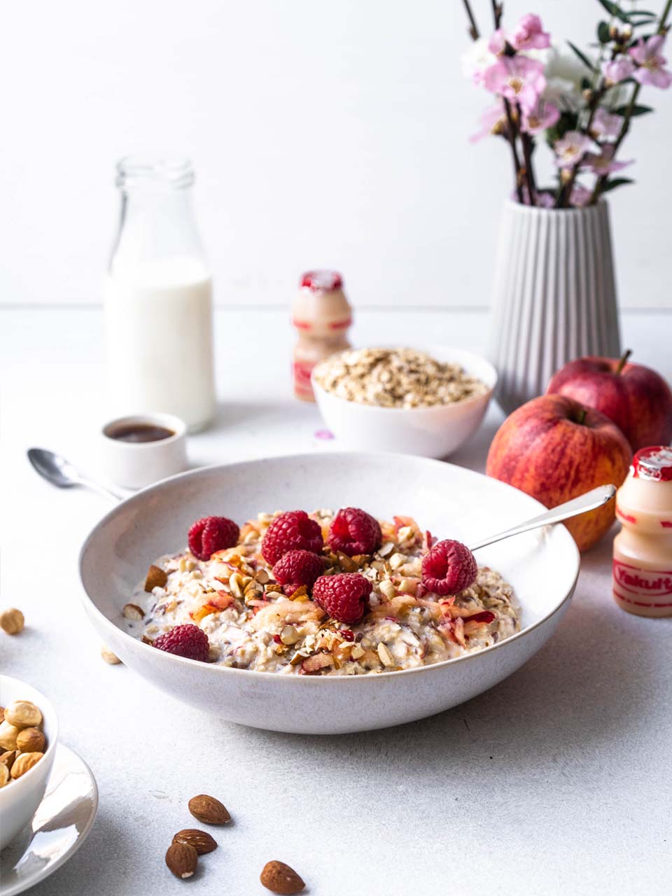 Muesli met frambozen in een kom, fles melk, kopje koffie,appels, noten, bloesem in een vaasje en een flesje Yakult Original.