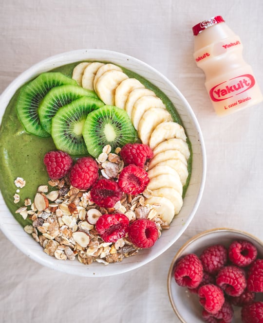 Een ontbijttafel met een kom met een groen kleurige bowl van een topping van kiwi, banaan, framboos en granen. Hiernaast staat nog een schaaltje met framboos en een Yakult Original