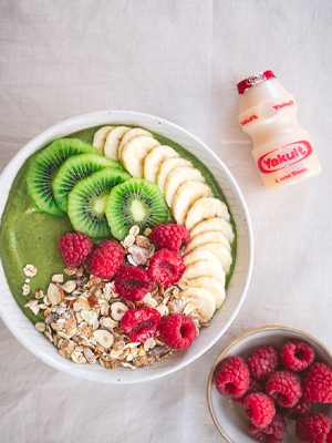 Een ontbijttafel met een kom met een groen kleurige bowl van een topping van kiwi, banaan, framboos en granen. Hiernaast staat nog een schaaltje met framboos en een Yakult Original